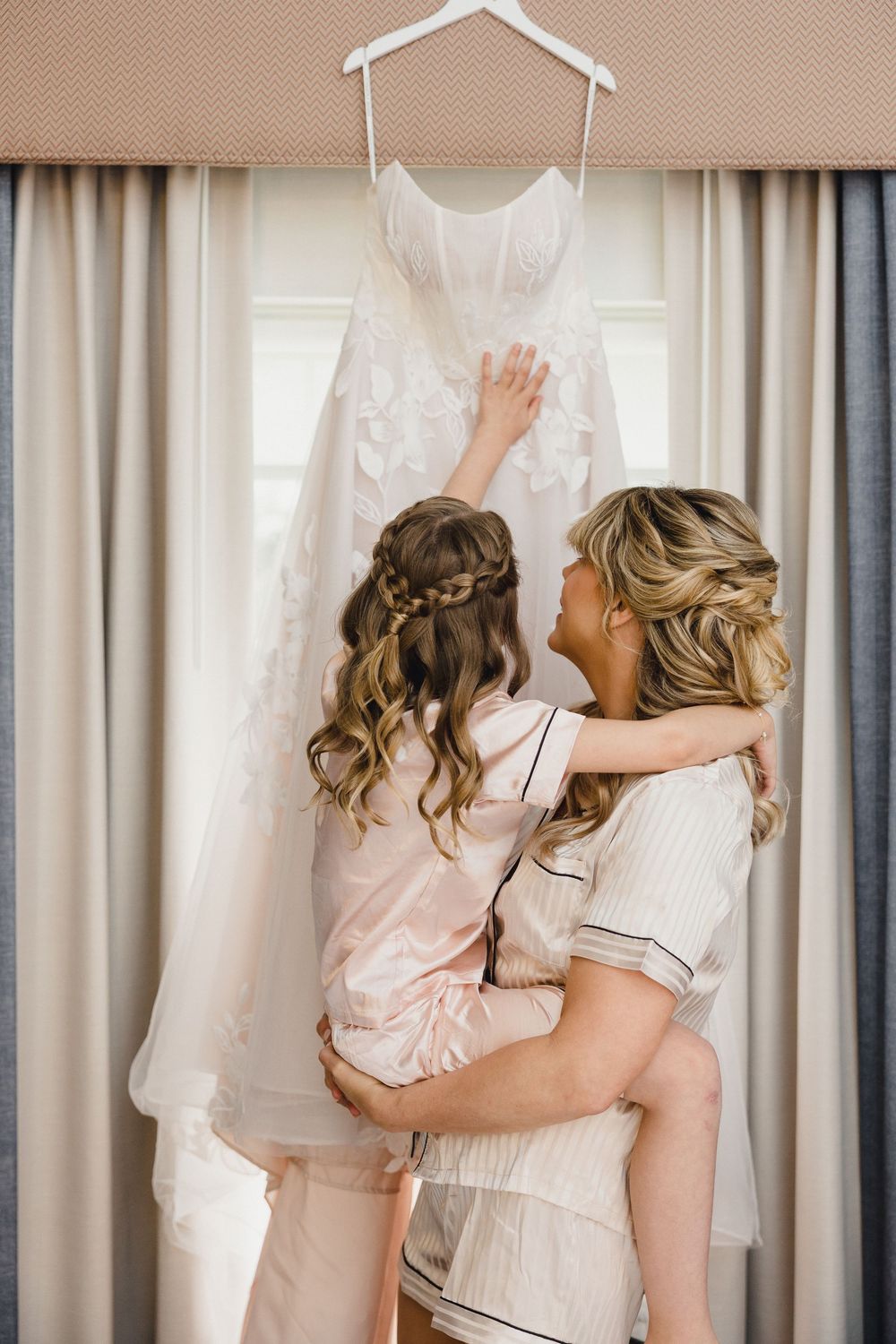 Women in pink robes examine white wedding dresses hanging on display in a bridal preparation room.