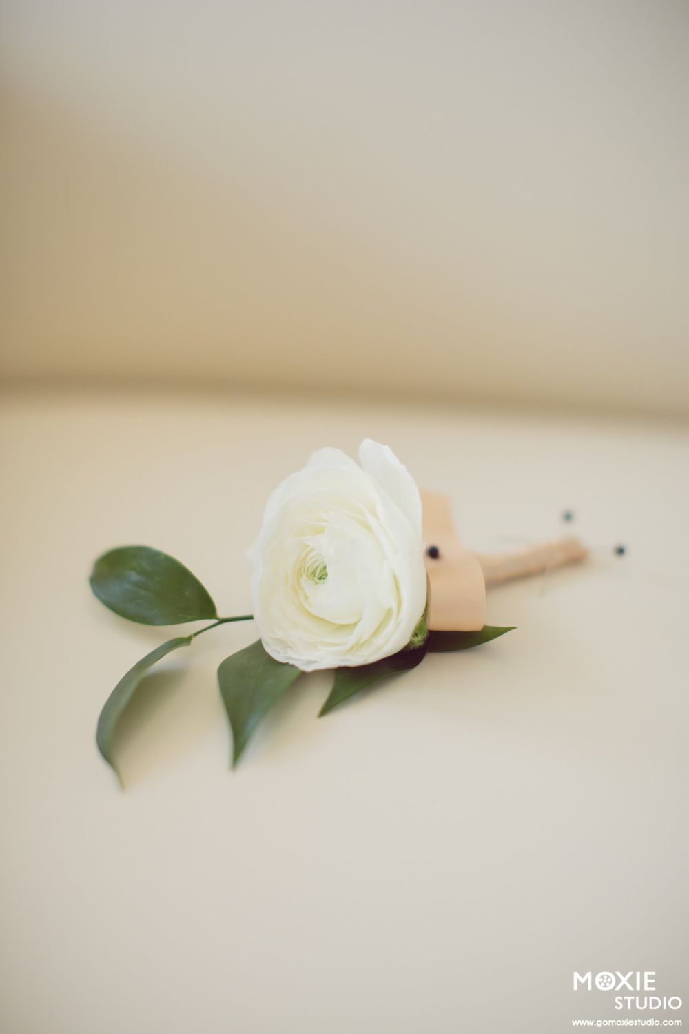 Single white rose boutonniere with green leaves against light background.