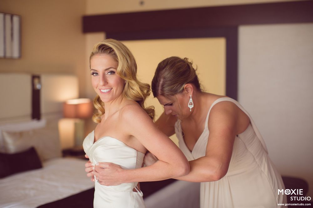 A bride gets help with her white wedding dress in a warmly lit hotel room.