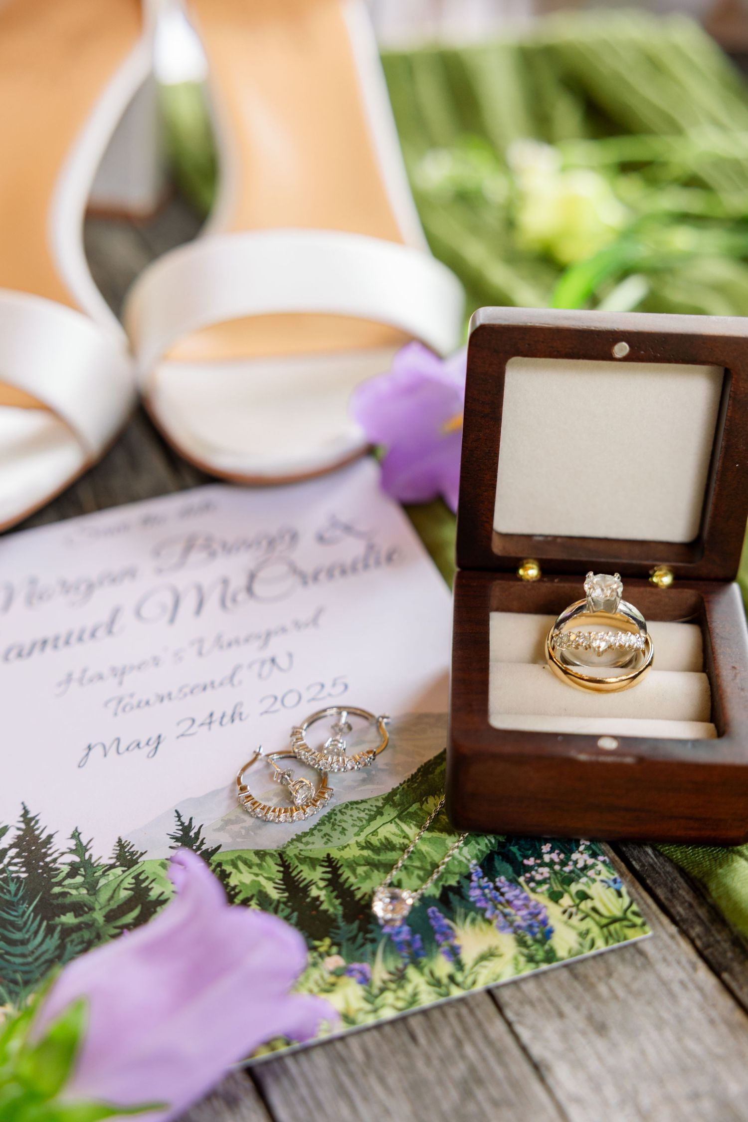 Wedding rings and jewelry displayed on rustic wooden surface with purple flowers and greenery decorations.