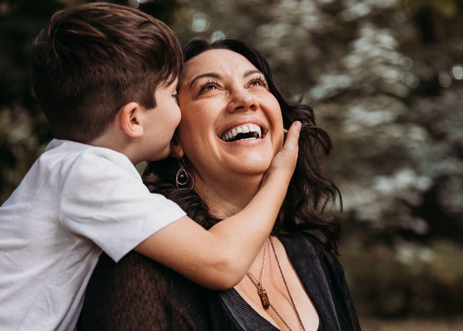 A child in a white shirt shares a tender moment with someone, both laughing joyfully outdoors.