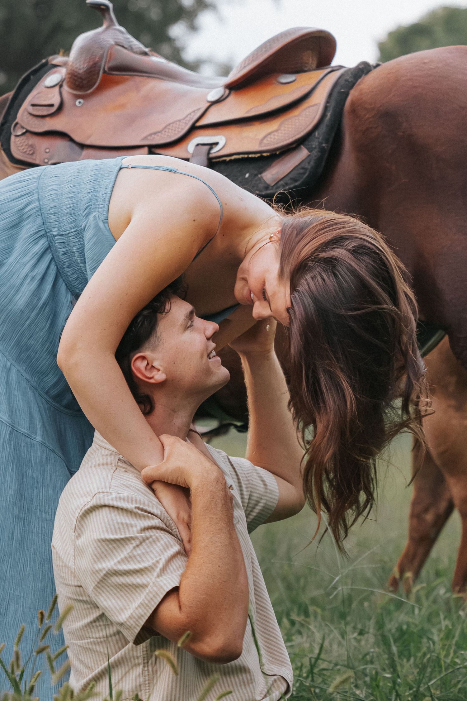 A romantic couple embraces in a field next to a saddled horse on a sunny afternoon.