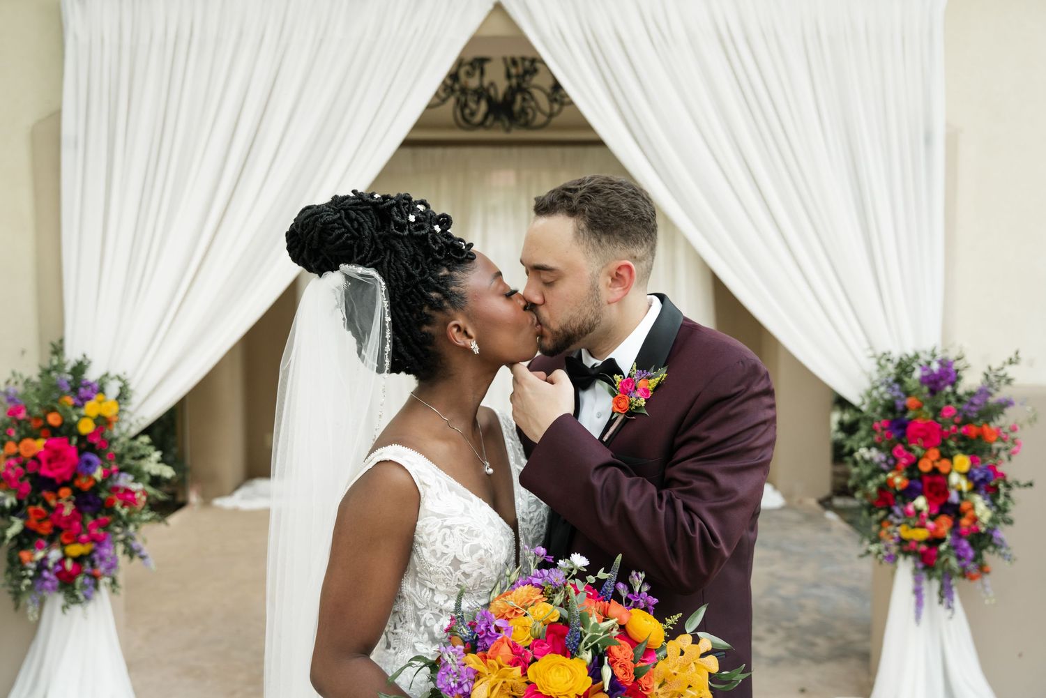 Wedding couple shares romantic kisses in a garden setting with vibrant floral arrangements and white drapery.