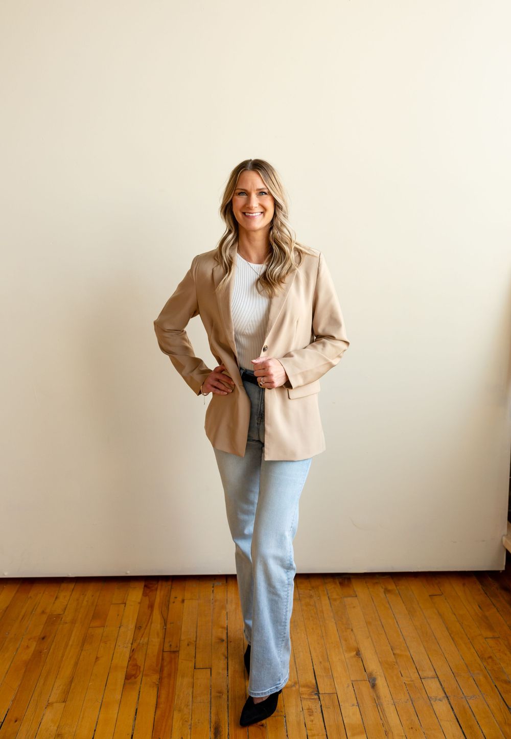 Professional wearing light beige blazer and blue jeans poses against white wall with hardwood floors.