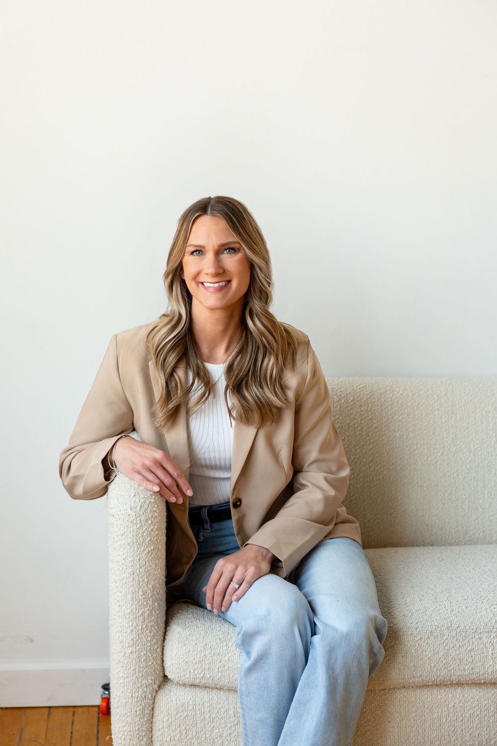 A person wearing a beige blazer and light jeans sits casually on a cream-colored couch in a bright room.