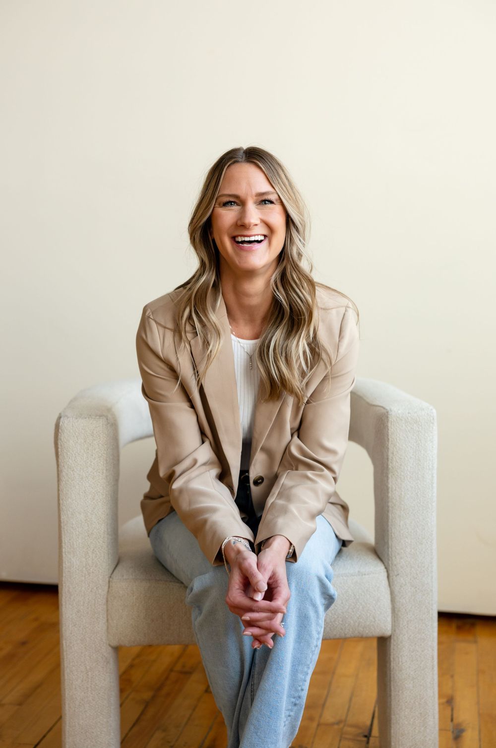 Professional lifestyle portrait of someone seated in a light gray armchair wearing beige blazer and jeans against white wall.