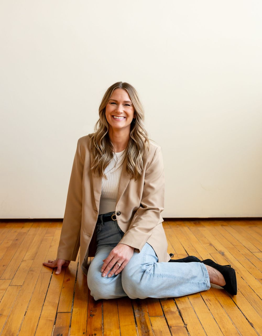 A series of casual poses on hardwood floor wearing a tan blazer, light sweater, and jeans.