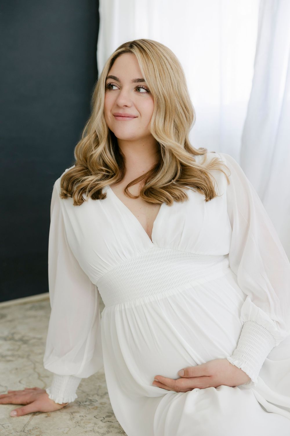 A pregnant woman in a flowing white dress sits by a window with soft natural lighting in an elegant maternity photo.