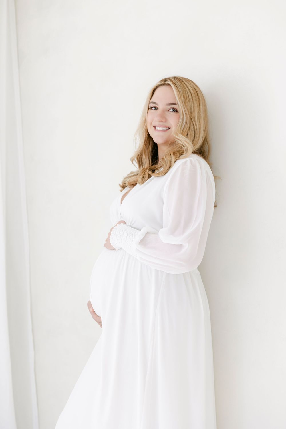 A pregnant woman in a flowing white dress stands against a white wall, gently holding her baby bump.