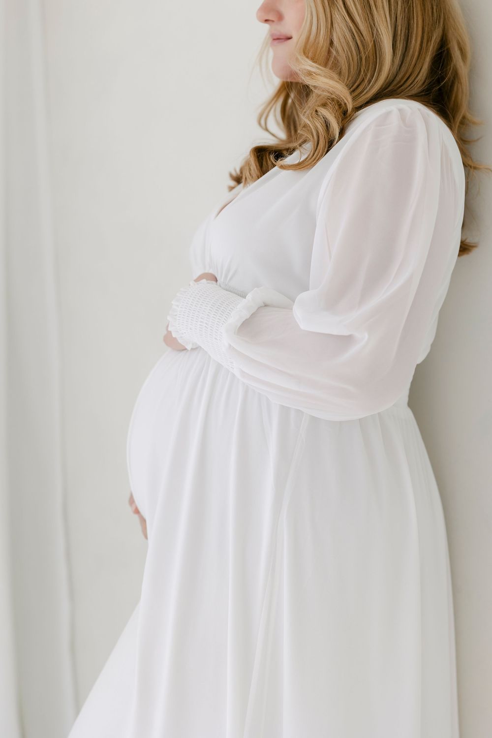 Side view of an elegant white maternity dress with sheer long sleeves and empire waist shown on an expectant mother.