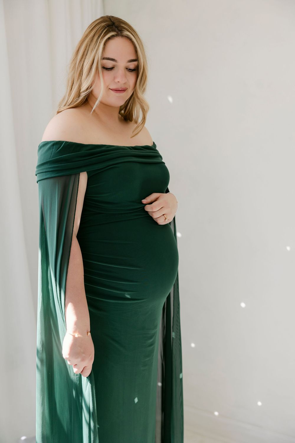 Elegant maternity portrait in emerald green off-shoulder gown with flowing cape sleeves against white backdrop.