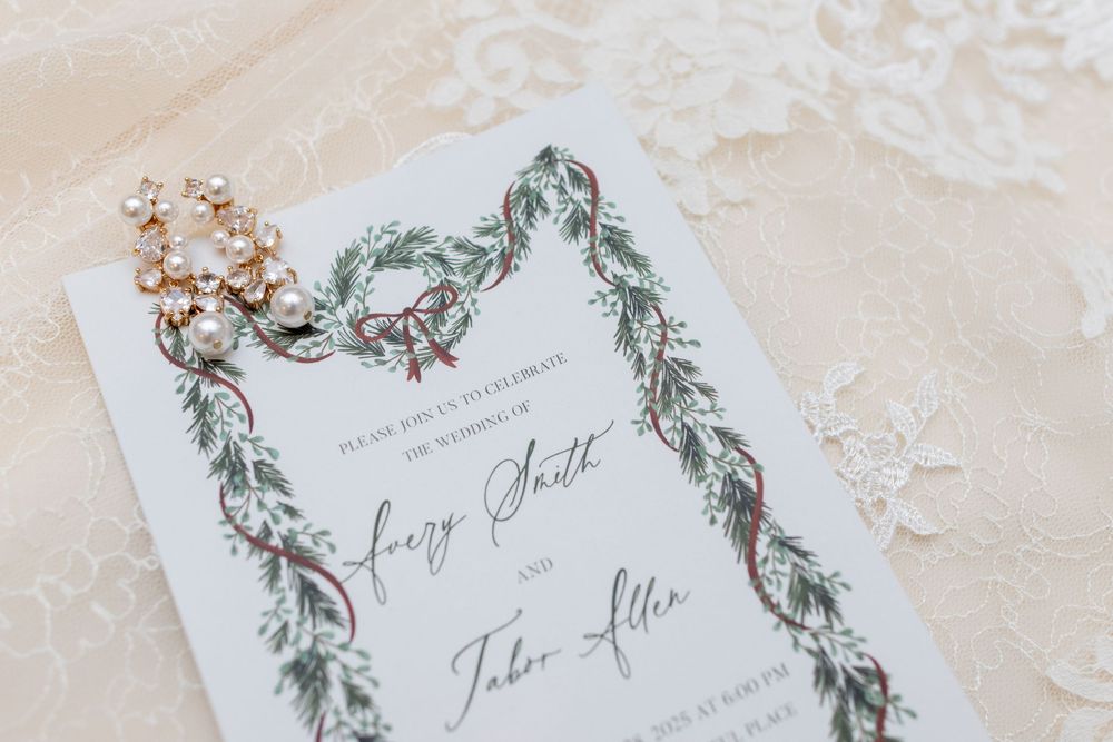 Elegant wedding invitations with sage green botanical designs and calligraphy text laid out on white lace fabric.