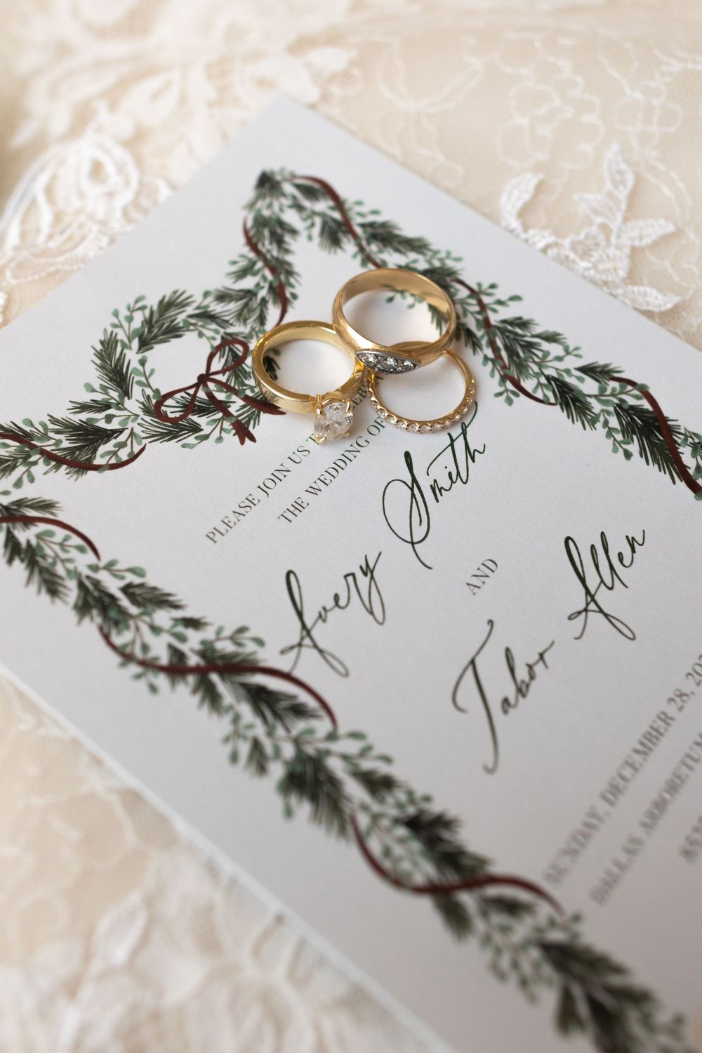 Wedding invitations with evergreen branch designs lay beside gold engagement rings on a textured cream surface.
