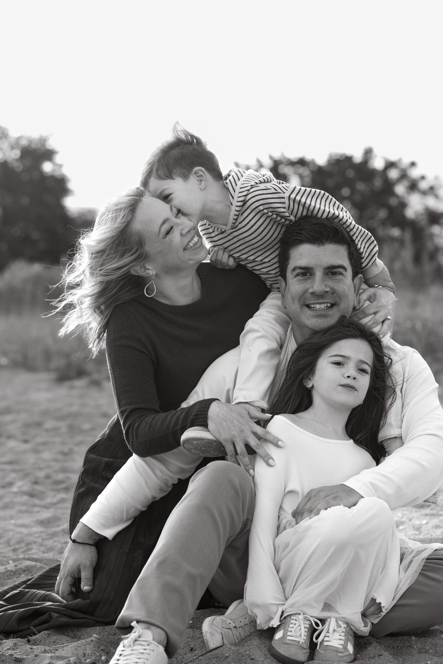 A family of four posing together on the sand during a sunset photoshoot at a Westport beach. Boy kissing mom's cheek.