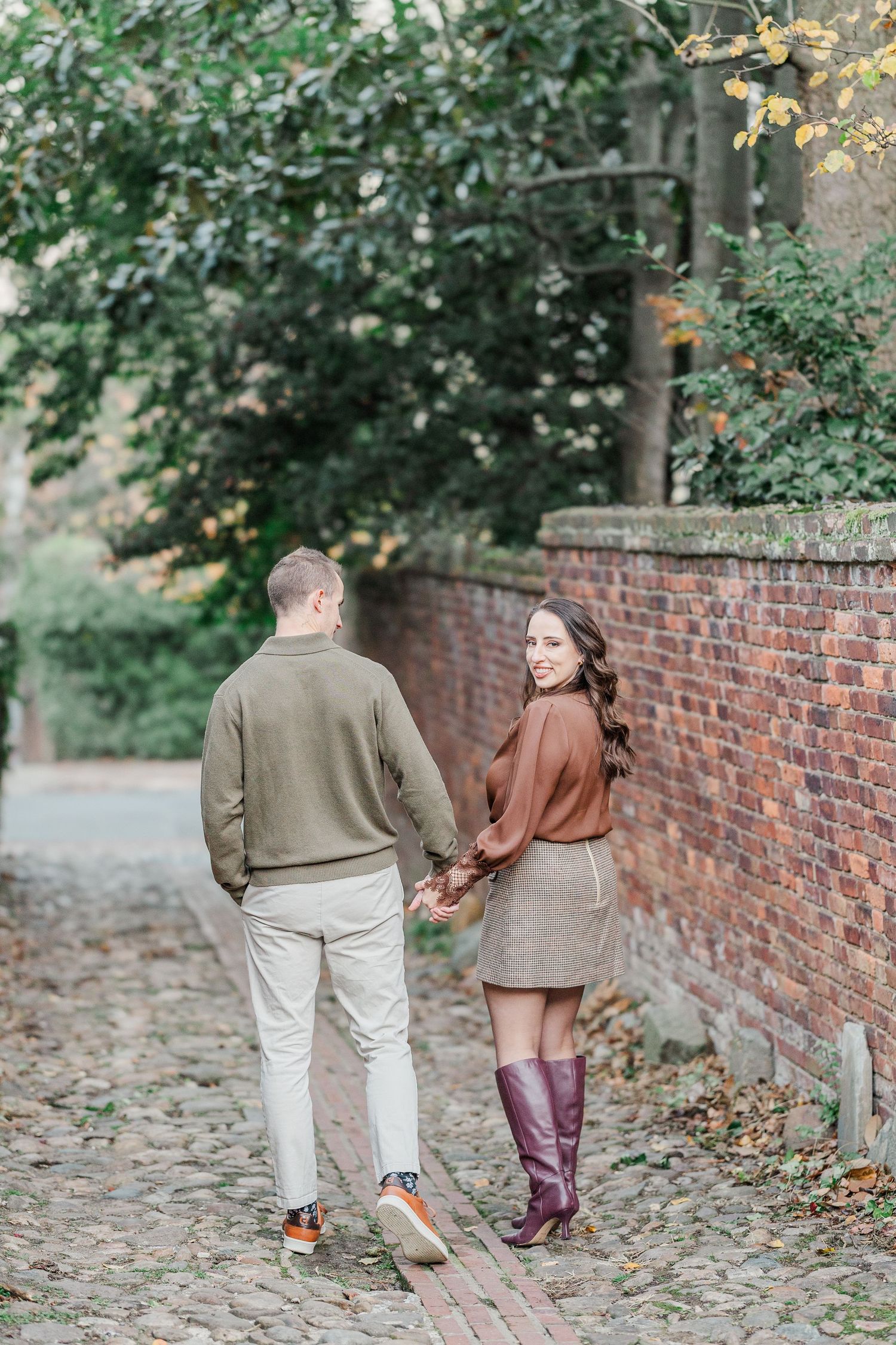 Couple walking together on a cobblestone path beside a brick wall during autumn with fallen leaves on the ground.
