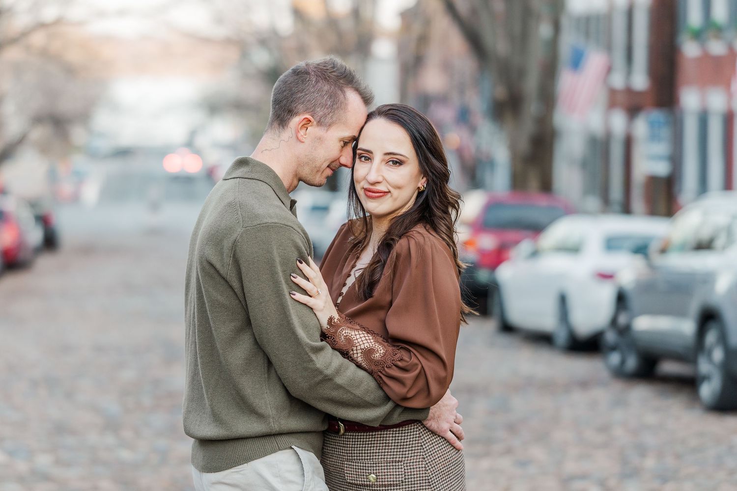 A couple embraces on a city street during autumn with parked cars and fallen leaves in the background.