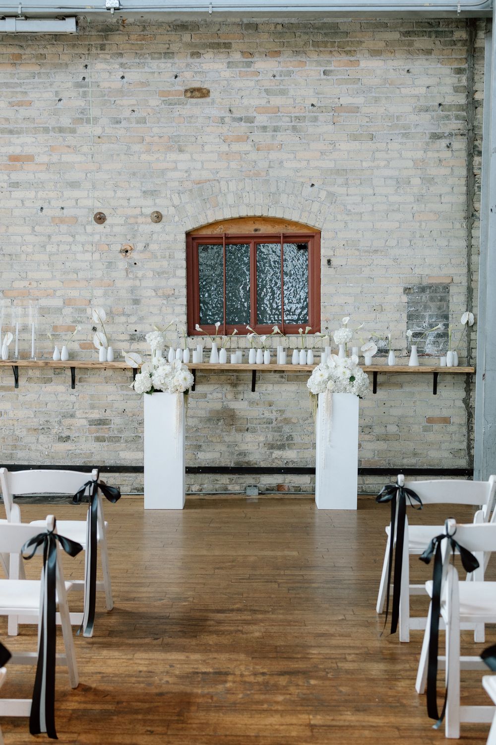 White floral centerpieces line long wooden tables in an industrial brick venue with dark curtains and hardwood floors.