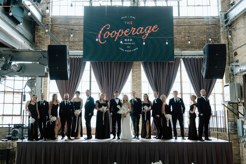Wedding party lined up in formal attire against industrial brick wall with vintage signage.