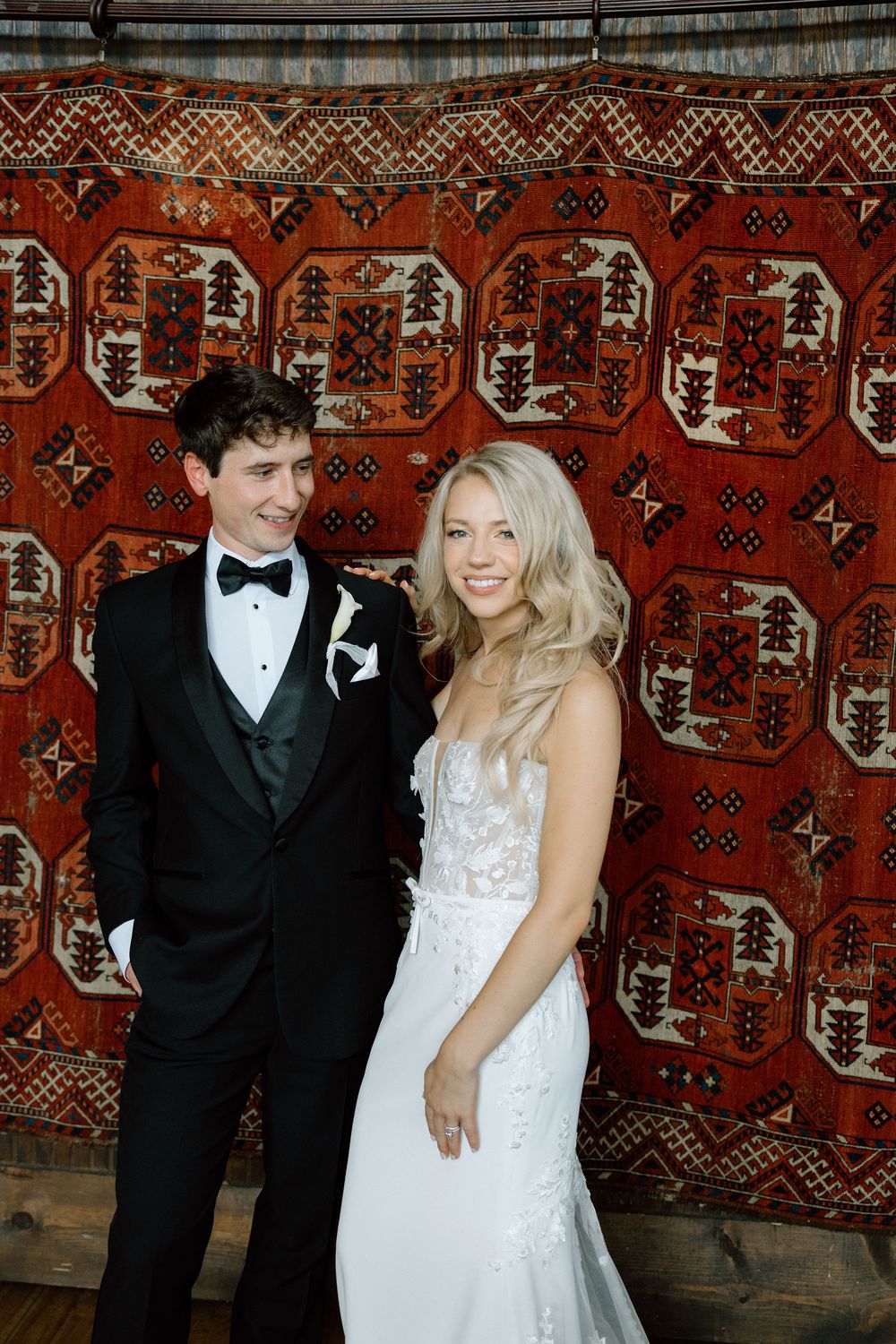 Wedding couple poses in front of decorative red patterned tapestry at reception venue.
