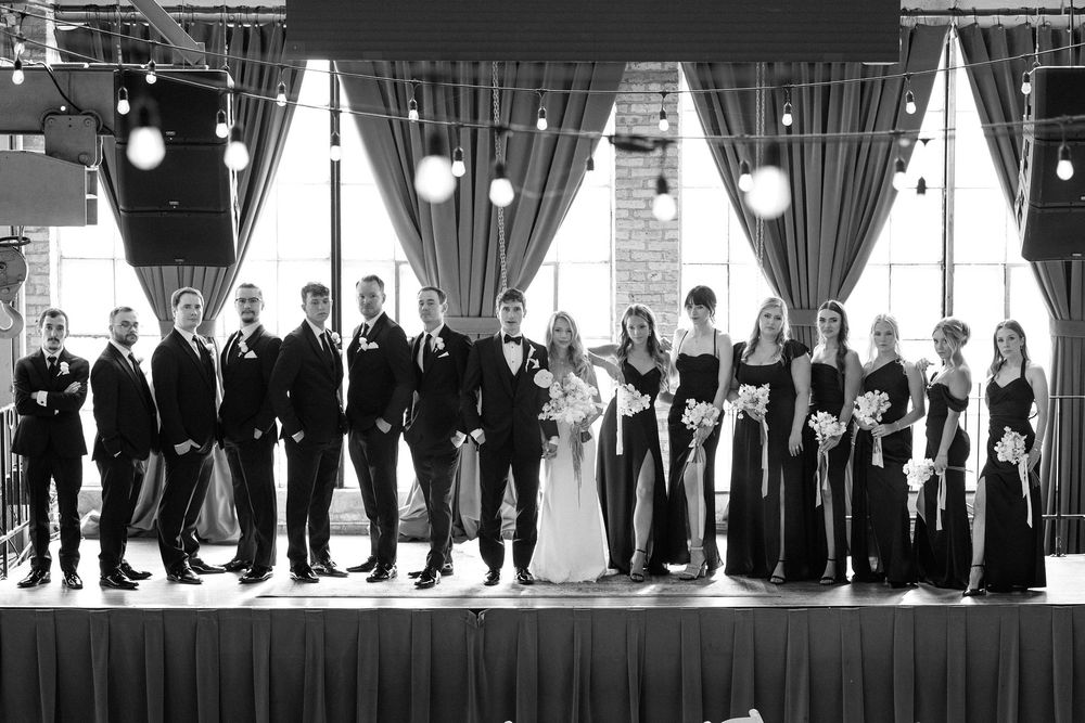 Black and white wedding party portrait showing full bridal party lineup against dark curtains.