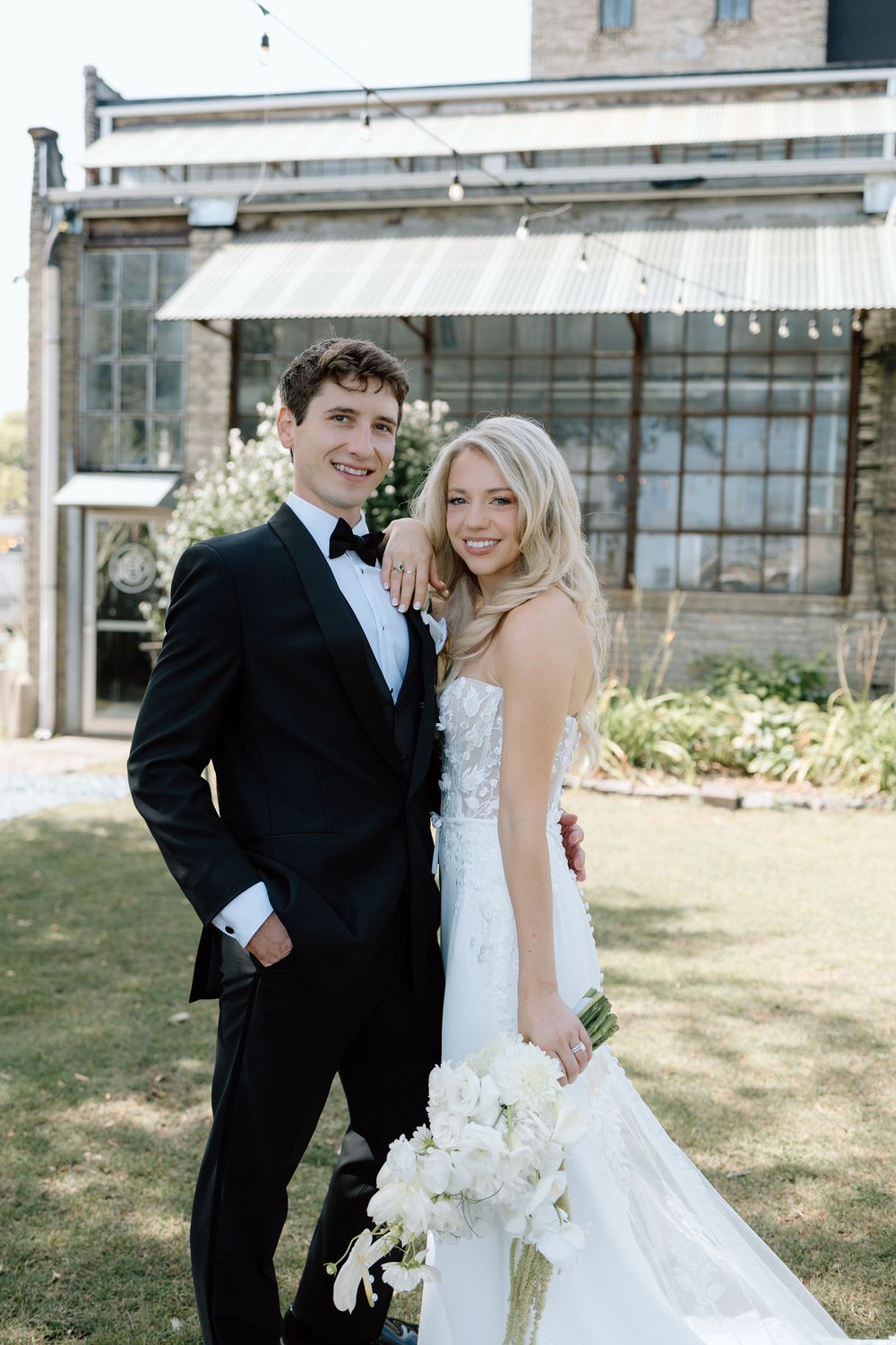 A bride and groom share joyful moments outdoors in formal wedding attire with a white floral bouquet.
