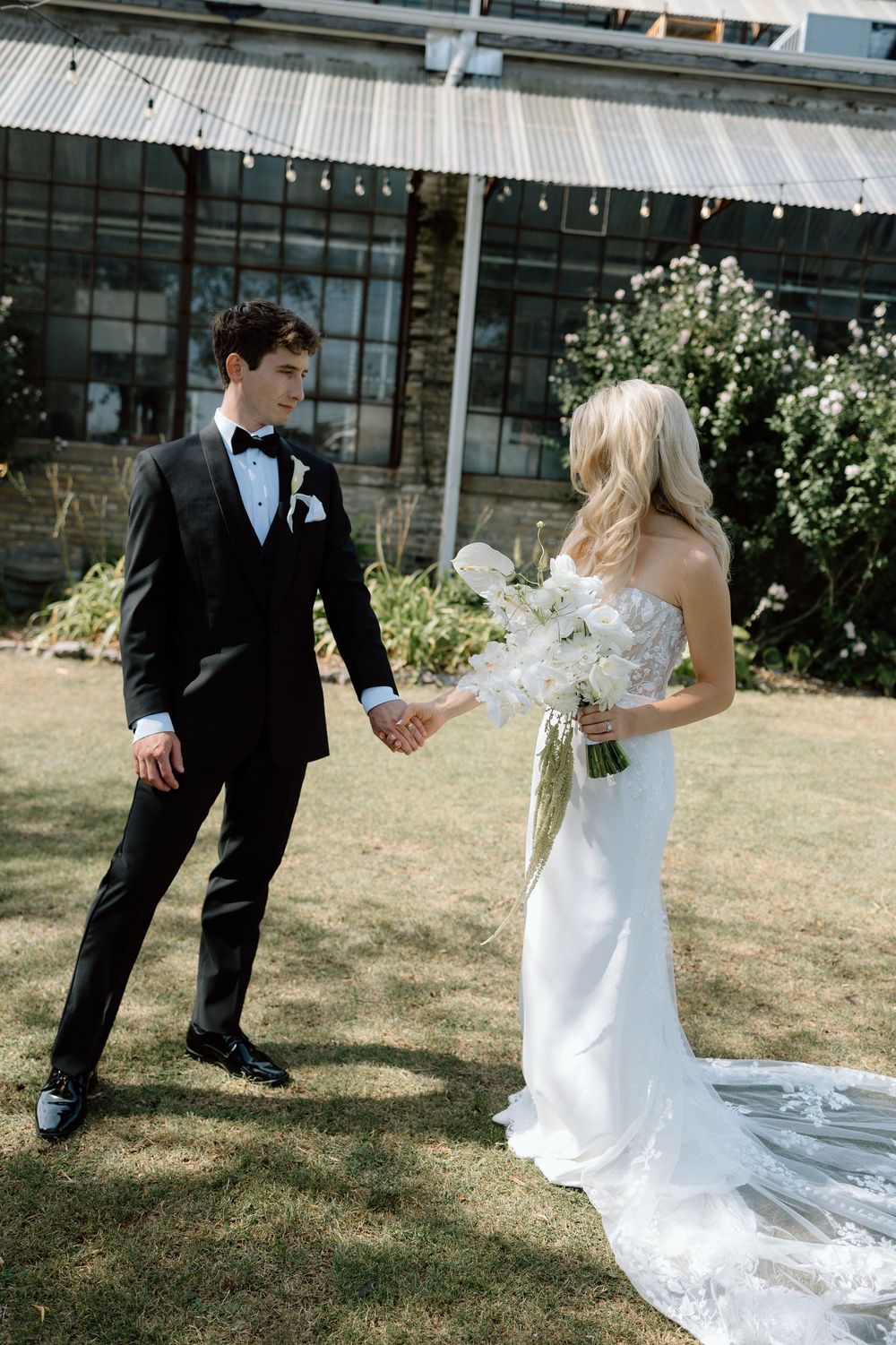 Wedding couple playfully interacts outdoors with flowing white dress and black tuxedo against rustic backdrop.