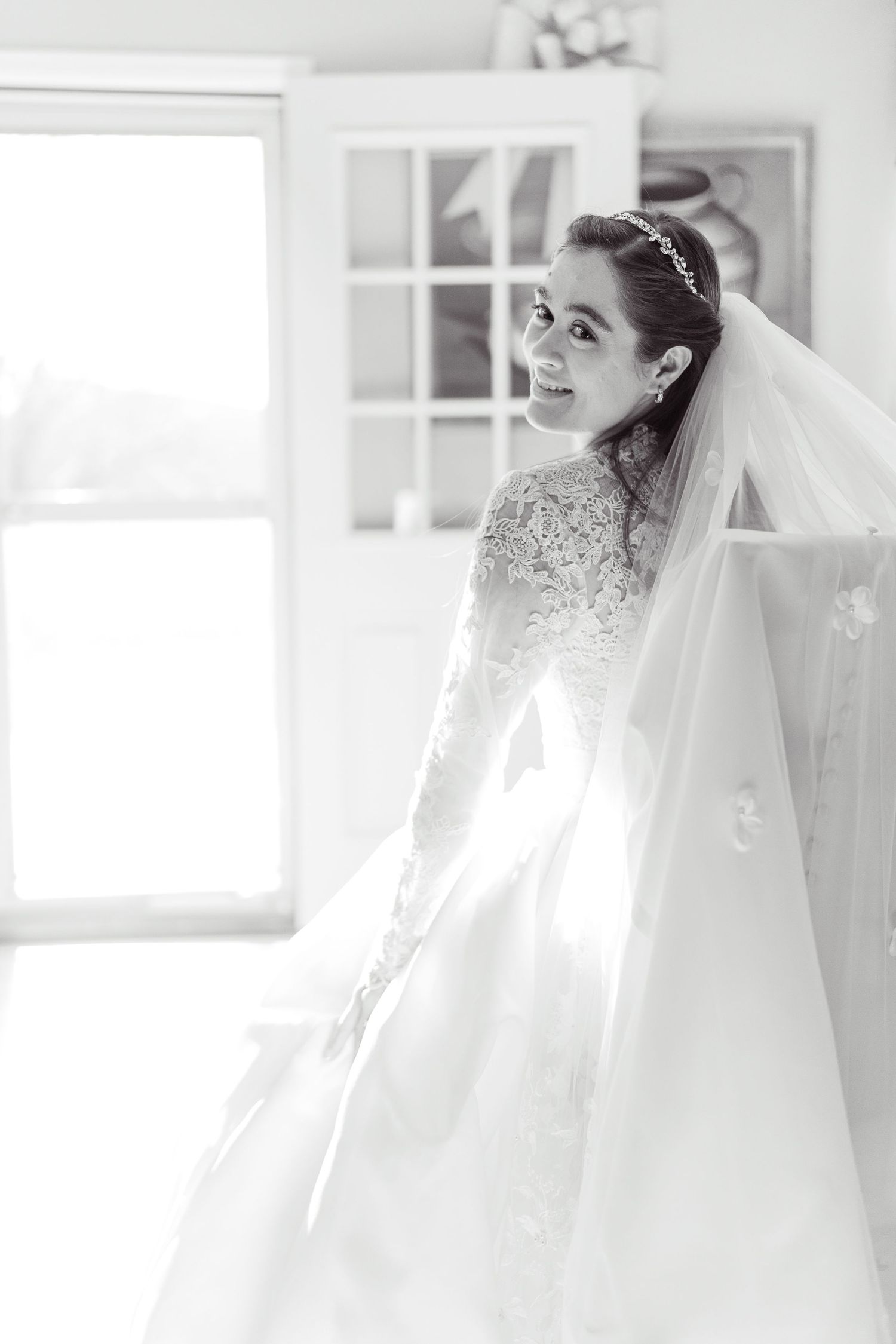 A bride wearing a flowing white veil and wedding dress smiles joyfully in a bright indoor setting.