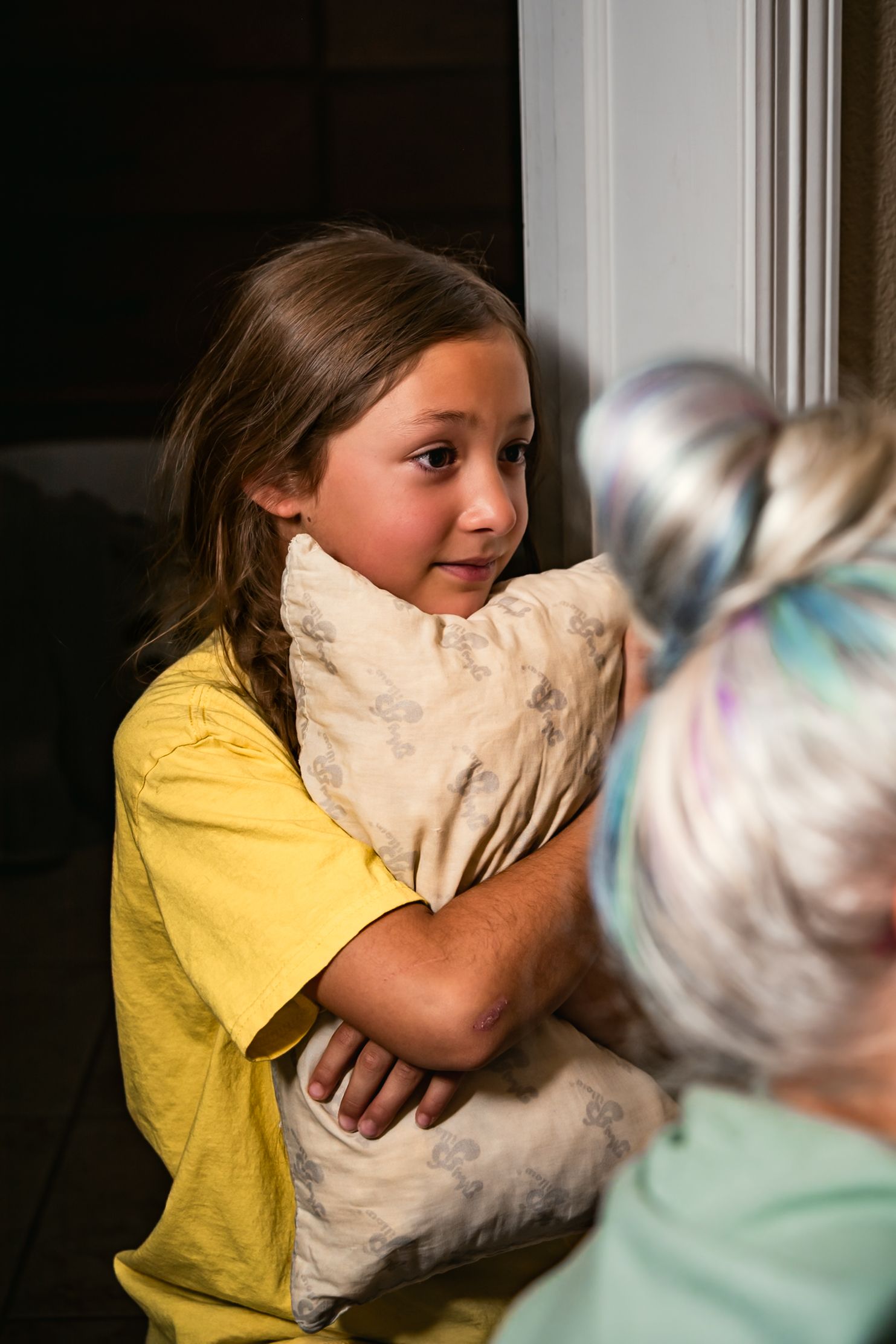 A big sister in a yellow shirt holds a pillow while looking on as her mother gives birth to her baby brother.