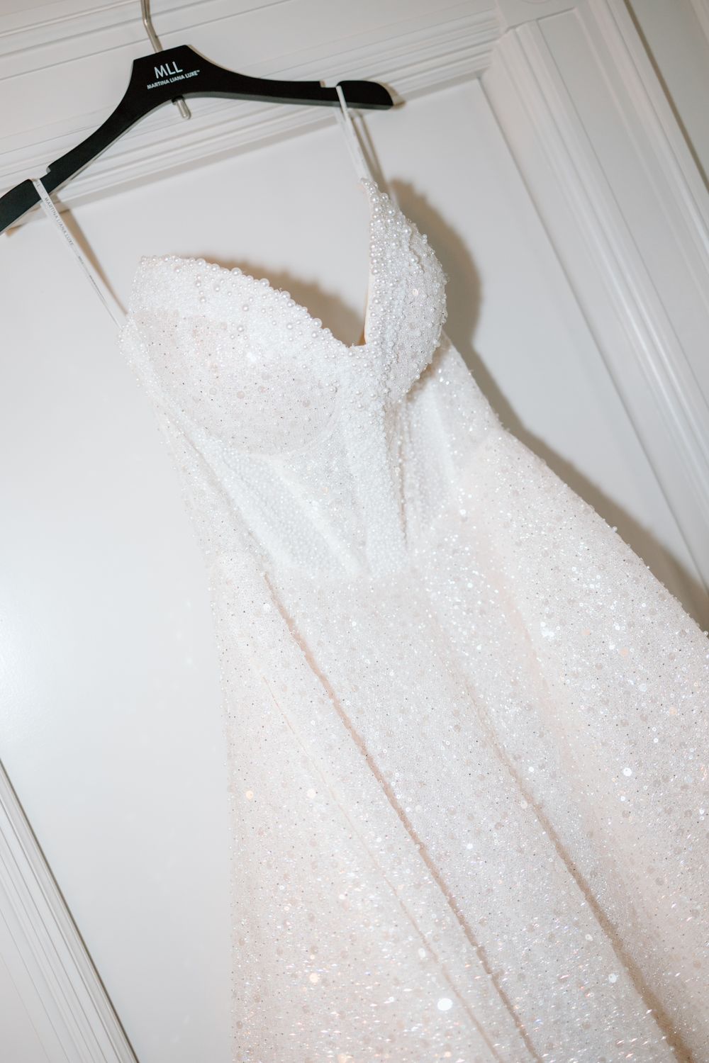 A sparkly white wedding dress with a sweetheart neckline and fitted bodice hangs on a black hanger against a white wall.