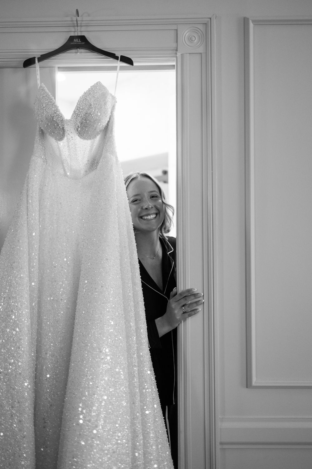 Sparkling white wedding dress hangs elegantly against white wall near window.