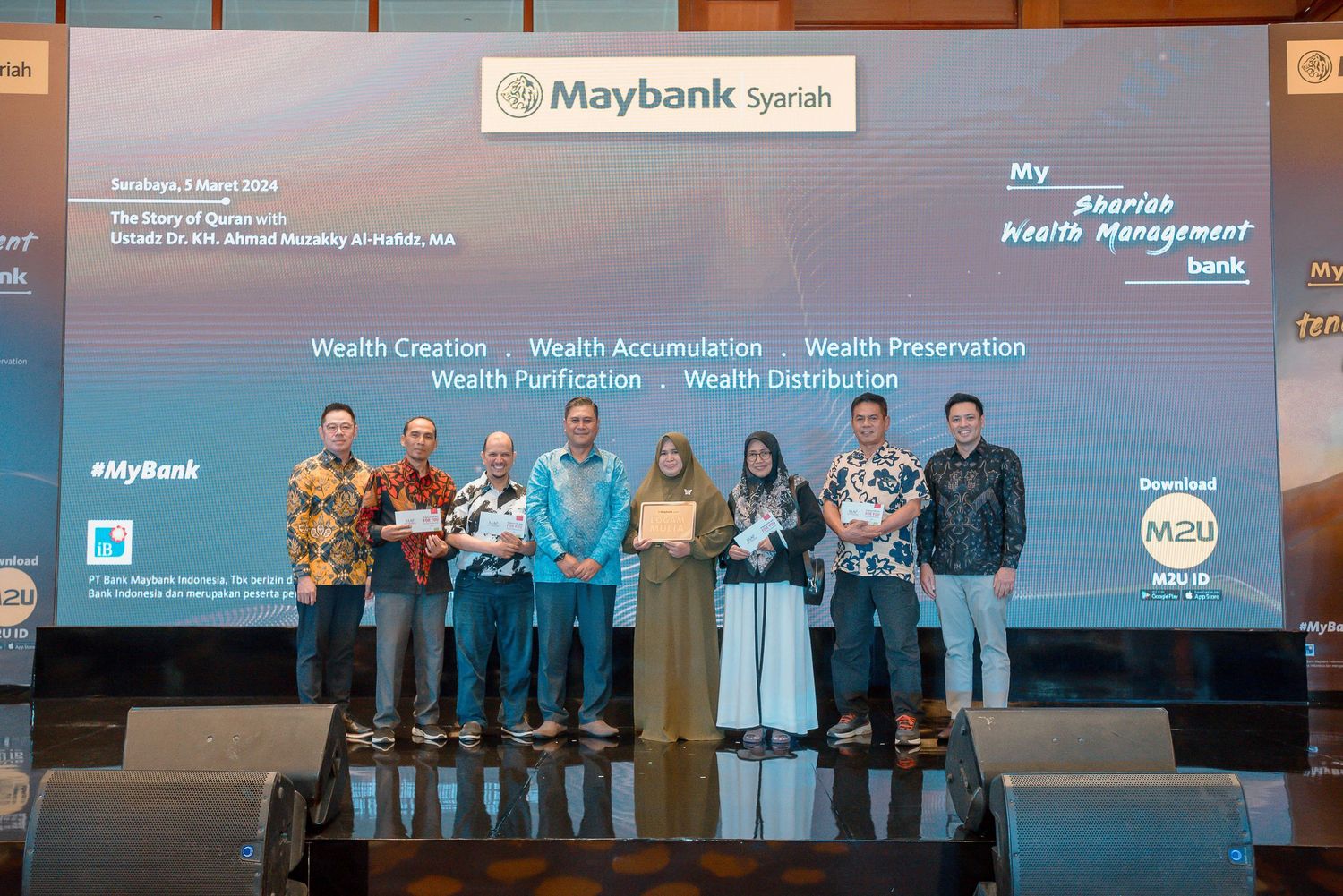 Business professionals stand on stage during a corporate Maybank event with presentation screens in the background.