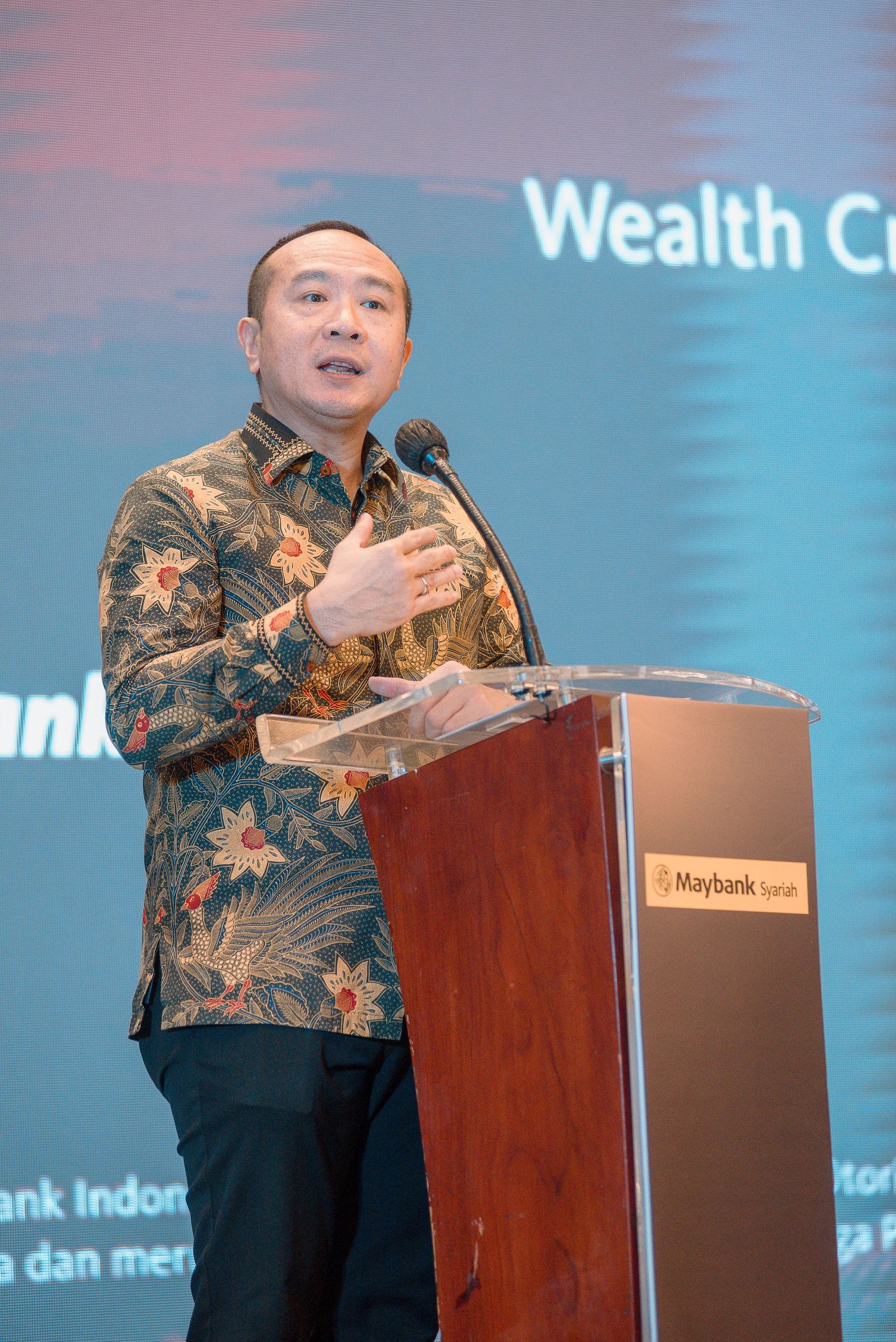 A speaker in a floral batik shirt delivers remarks at a podium during a Maybank wealth conference event.