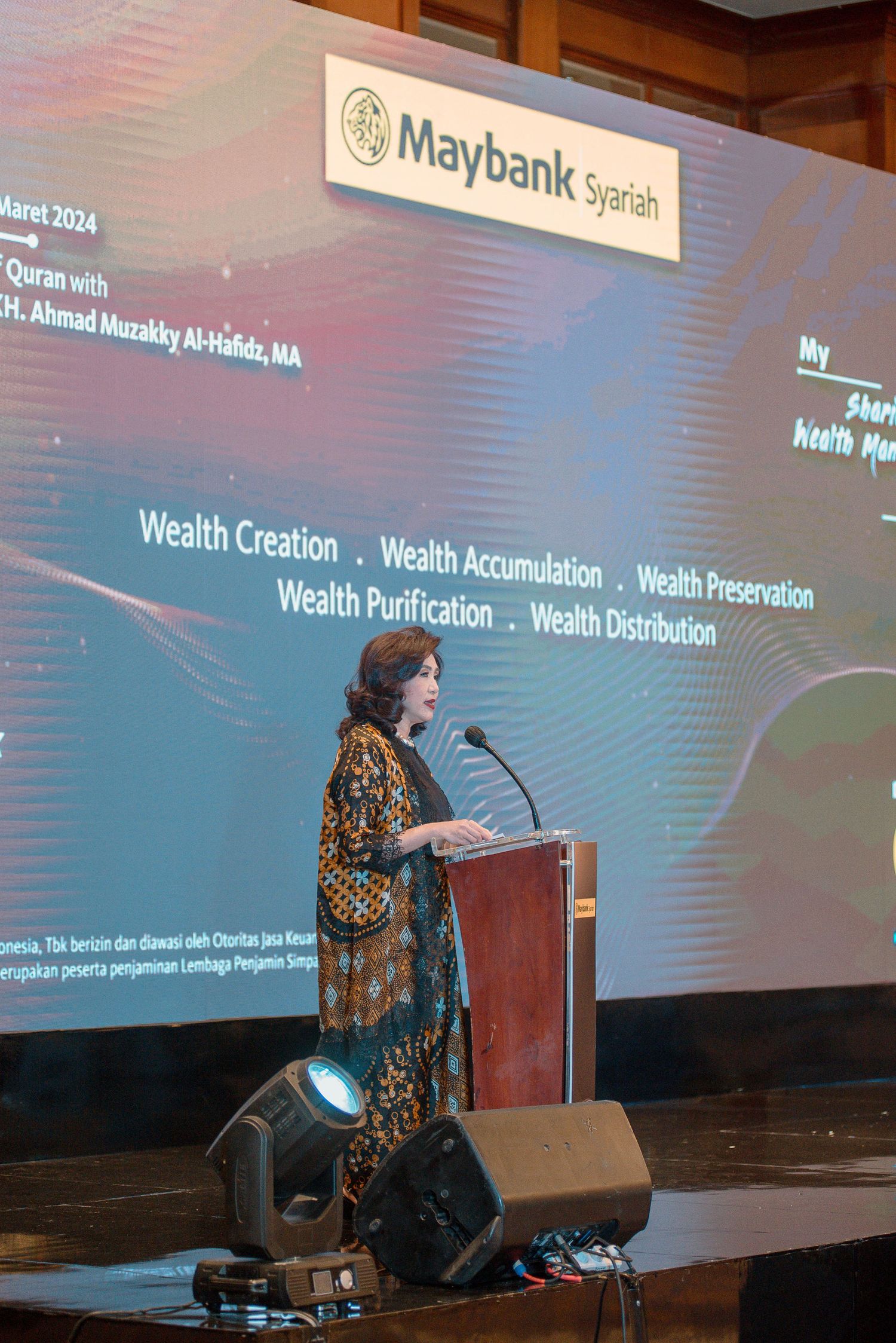 Speaker presenting at a conference podium with large projection screens showing wealth management topics in the background.