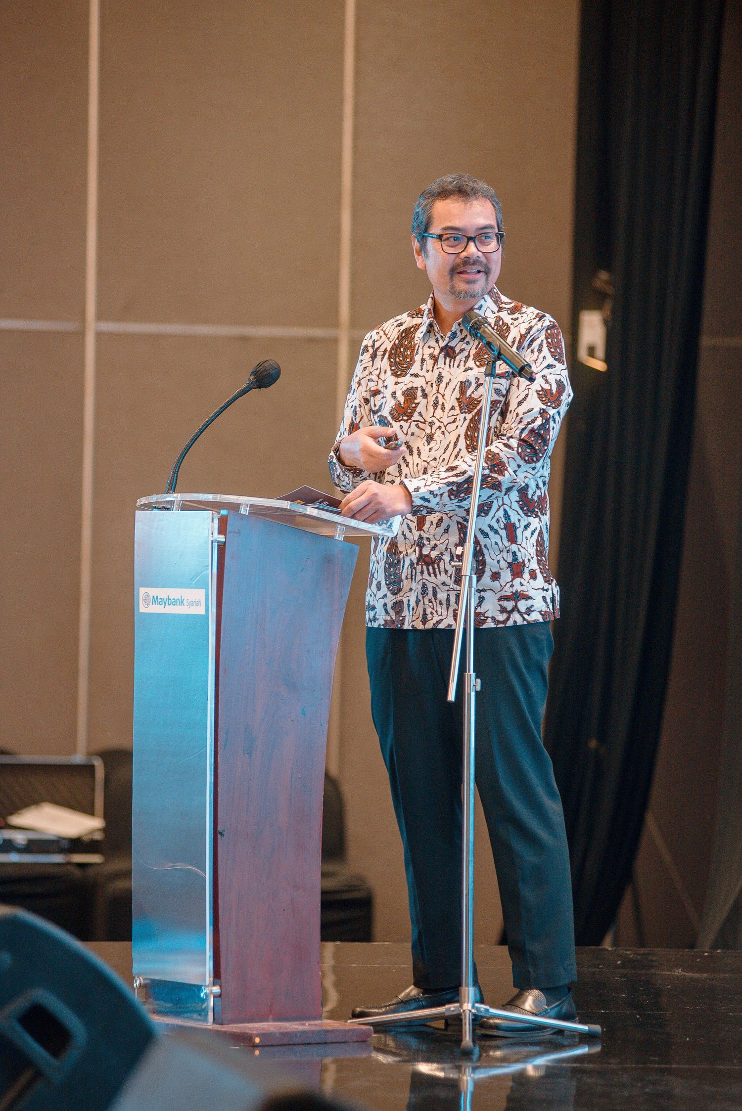 A speaker in a floral print shirt gestures while presenting at a podium during a conference presentation.