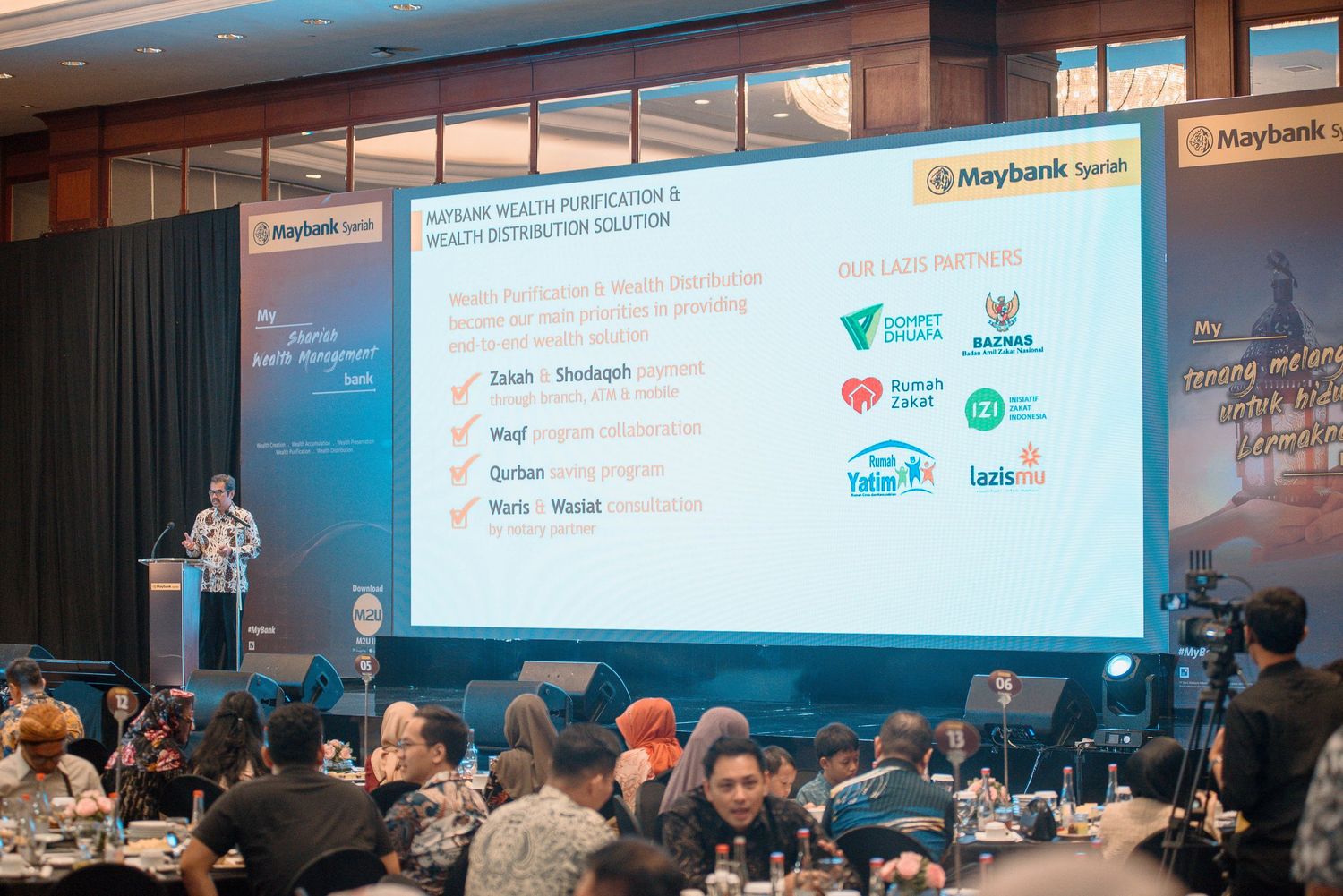 Business conference presentation showing sponsor logos on projector screen with audience seated at round tables.