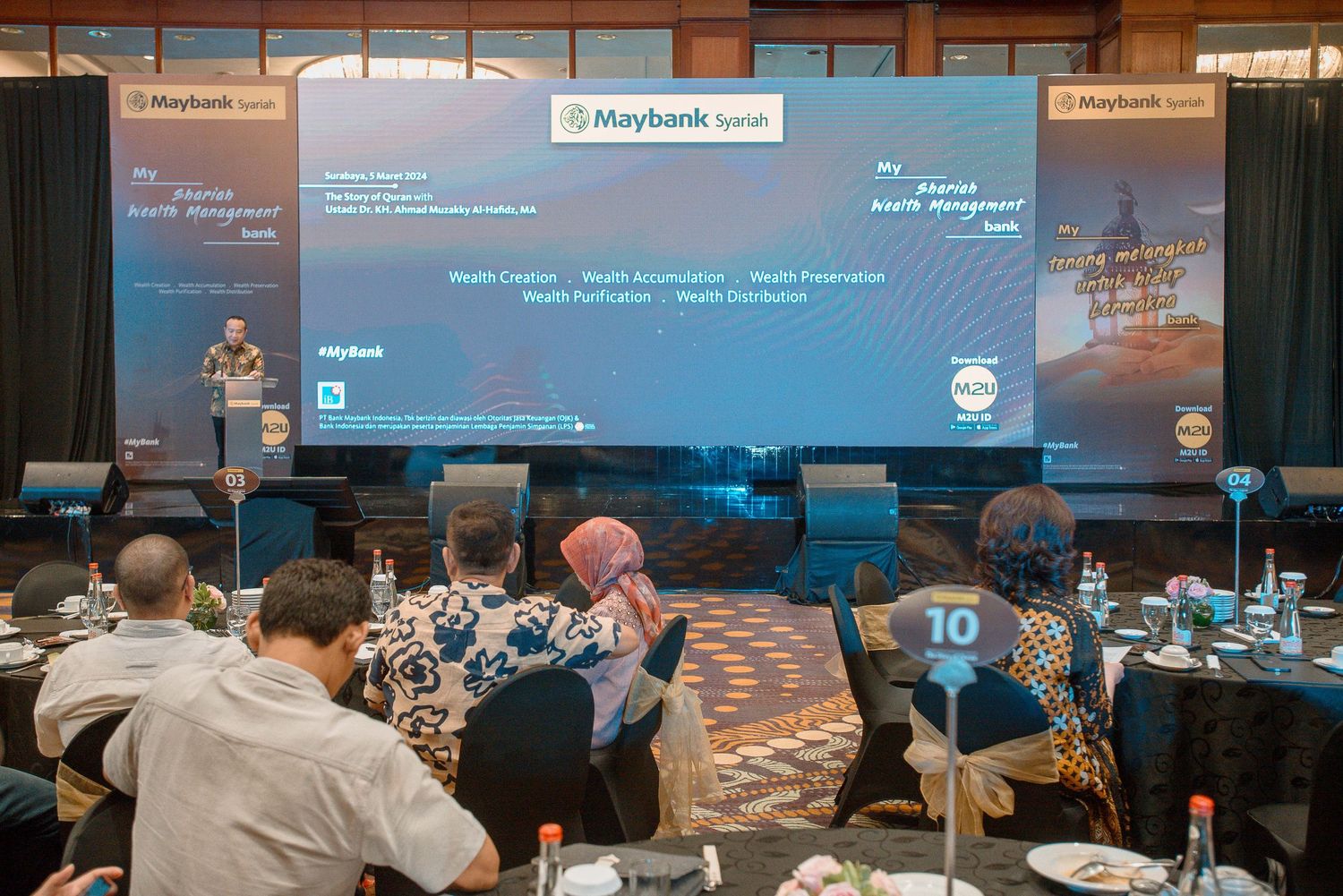 Conference attendees seated at round tables during a Maybank presentation with blue backdrop and digital displays.