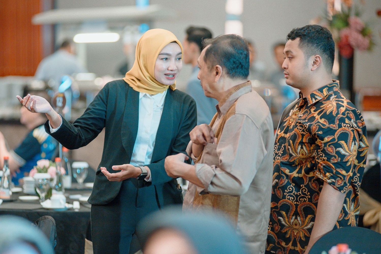 People having a conversation at a social gathering, with one wearing a yellow hijab and navy blazer and another in a batik shirt.