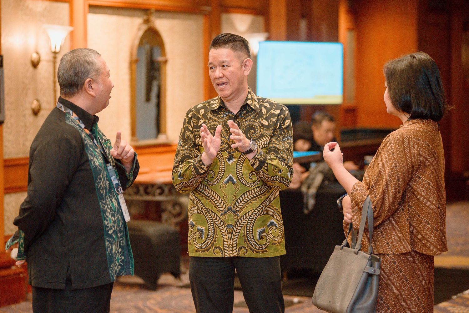 Three people in traditional batik attire engaged in animated conversation at an indoor business event.