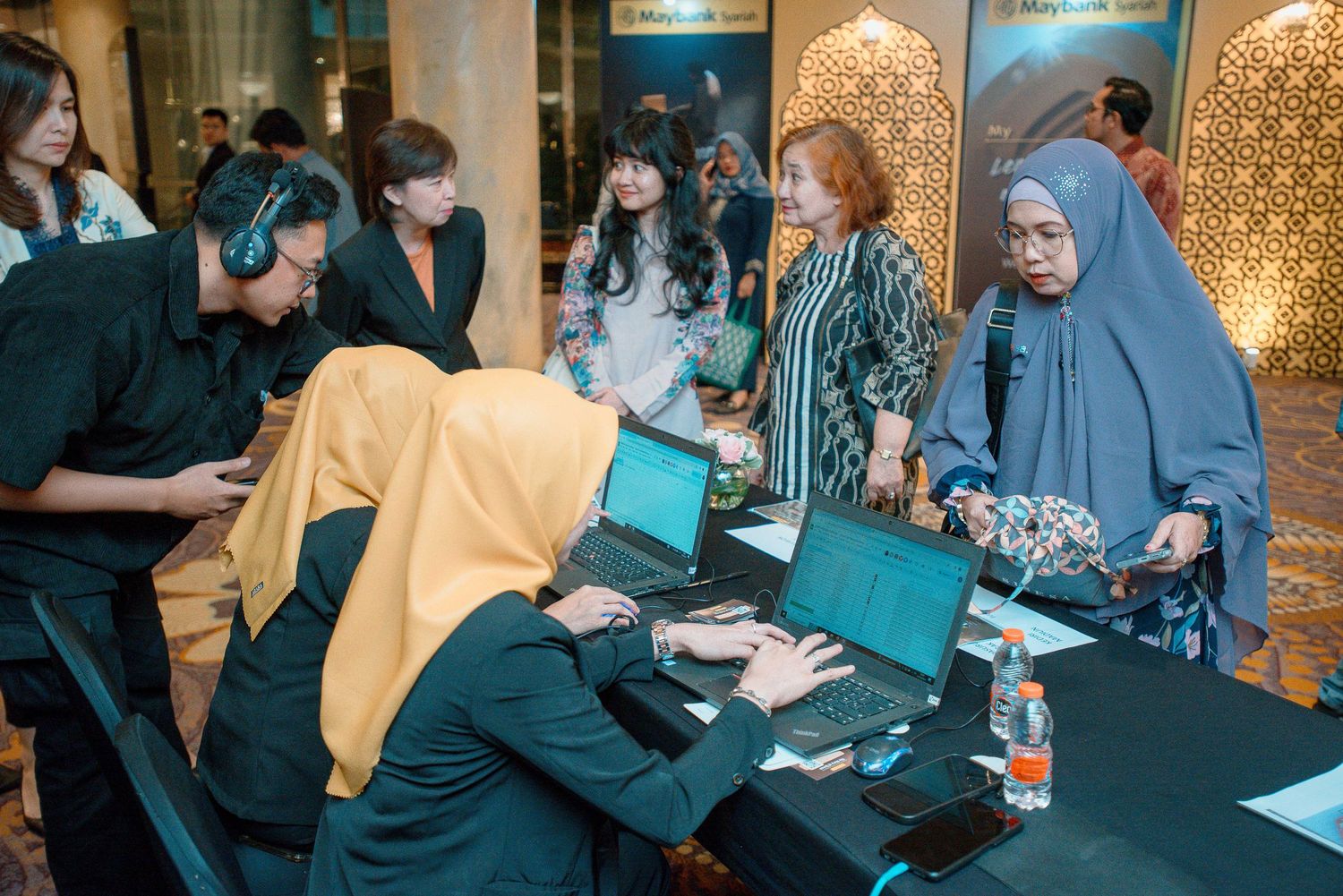 People sitting at tables signing documents and meeting with others in an office environment with decorative lighting.