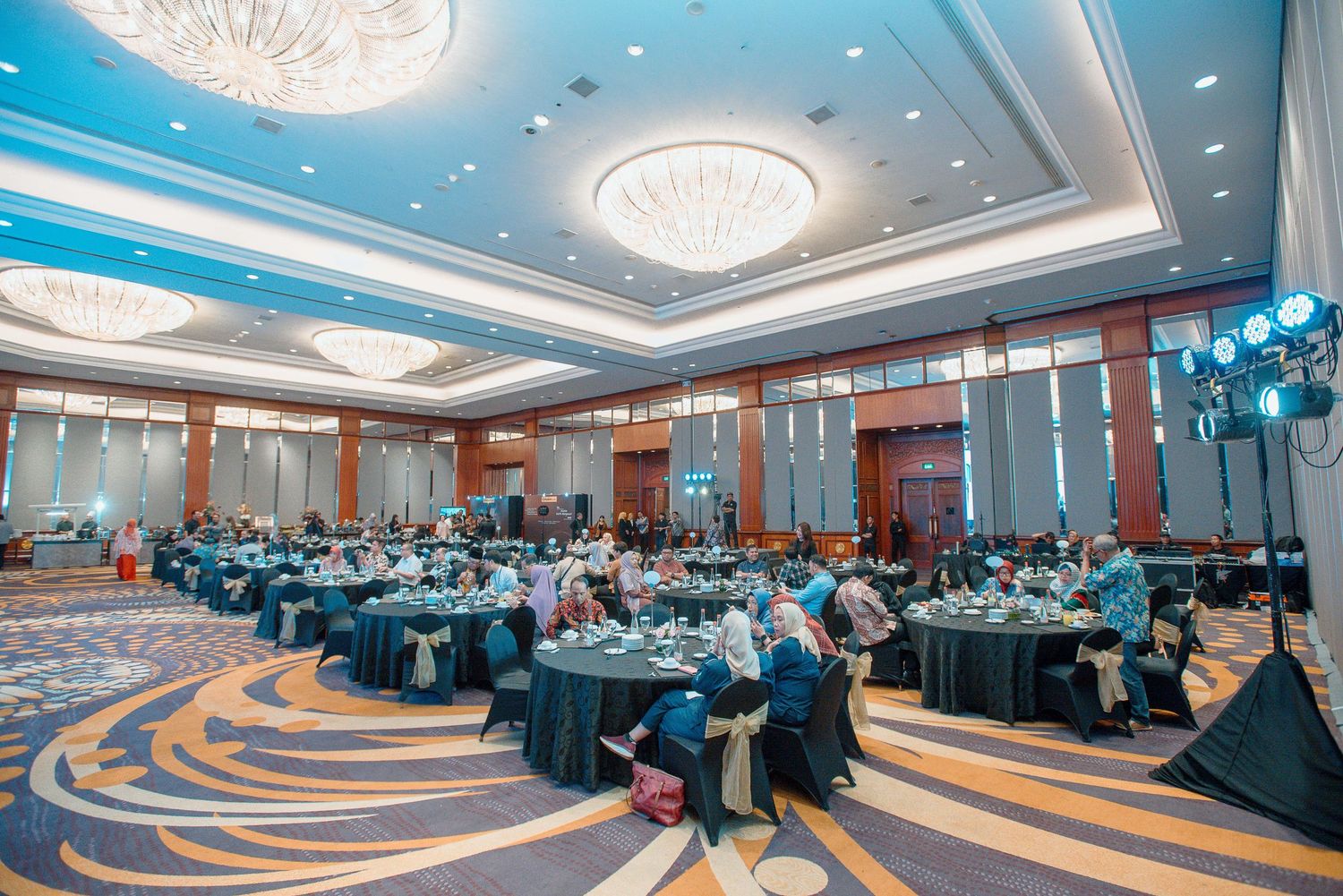 A panoramic view of a corporate conference room with round tables, chandeliers and attendees during a business event.