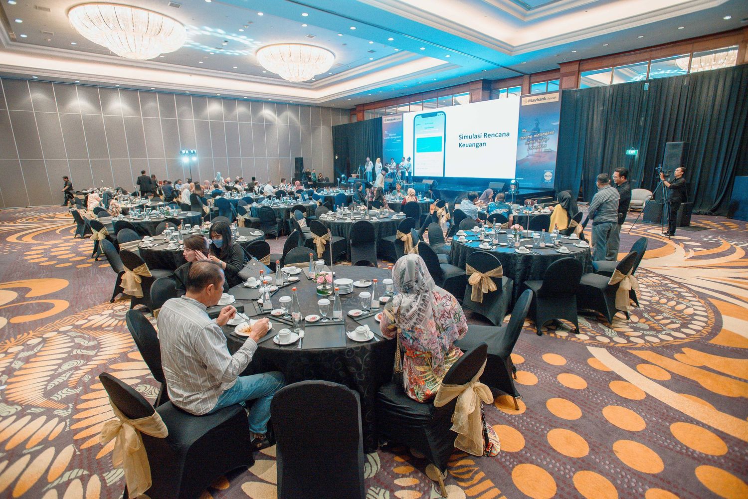 Conference attendees seated at round tables in a modern hotel ballroom with geometric carpet patterns and blue lighting.