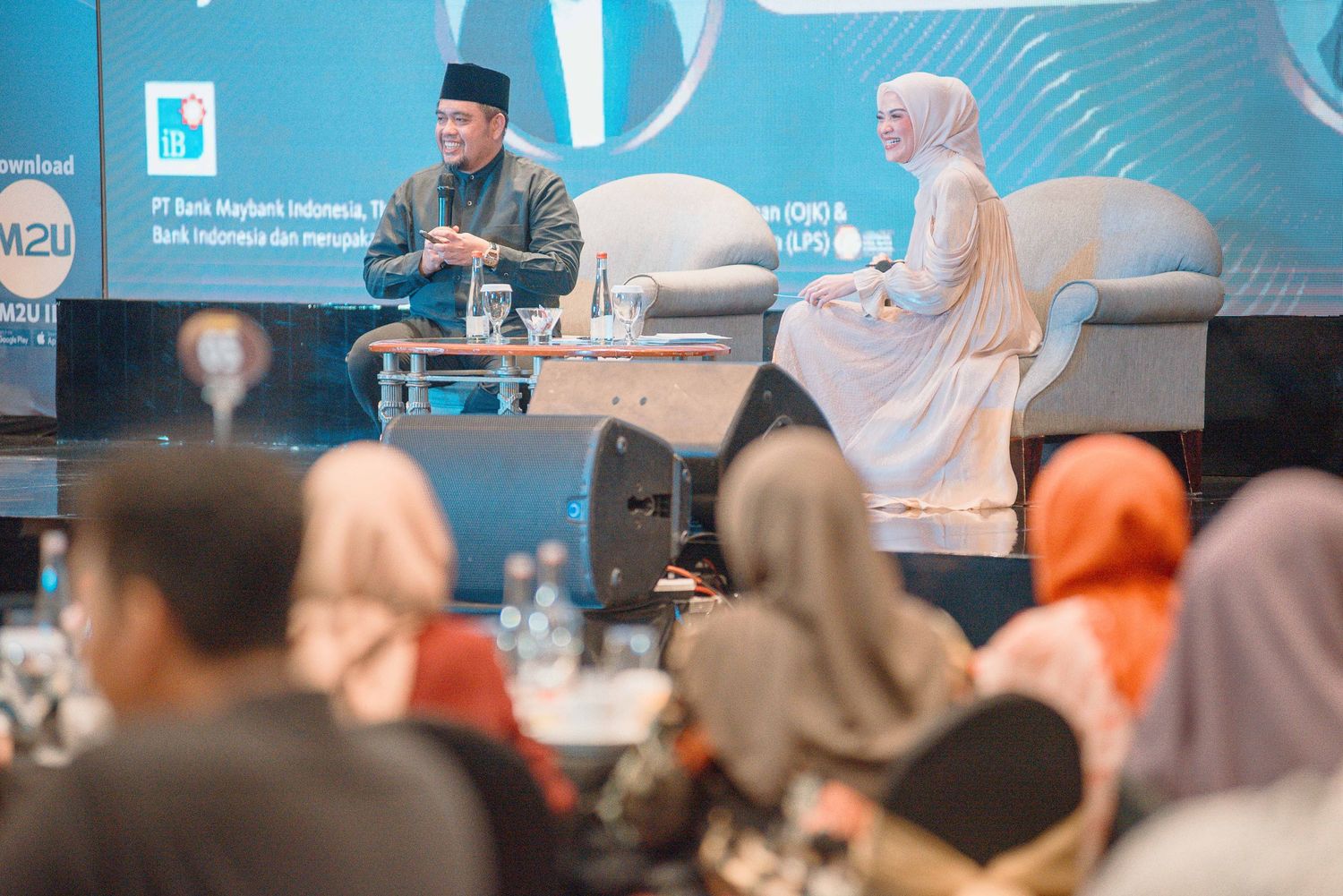 A speaker presents at a conference with audience members seated in rows listening attentively.
