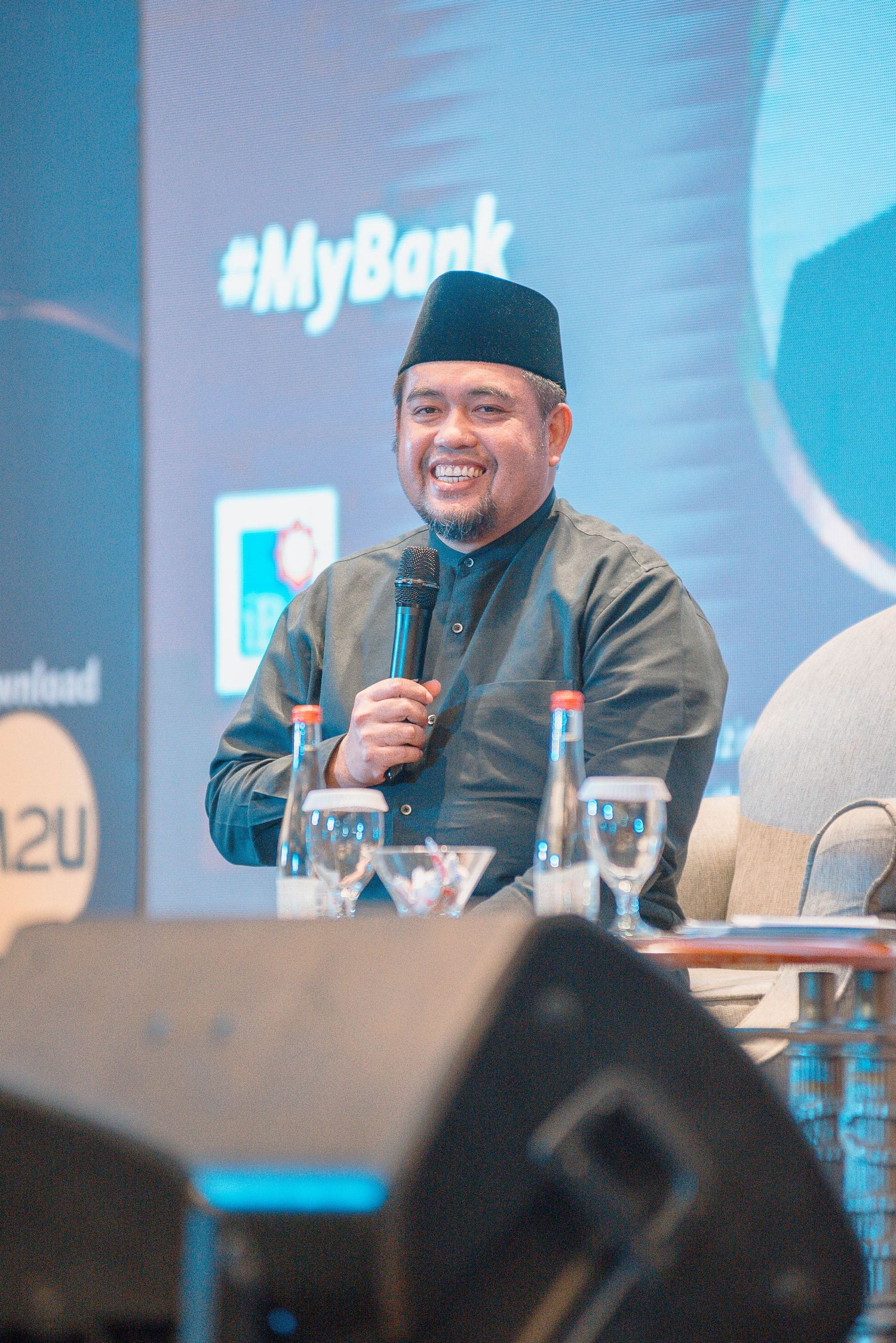 Speaker wearing traditional black cap smiles while participating in panel discussion at conference event.