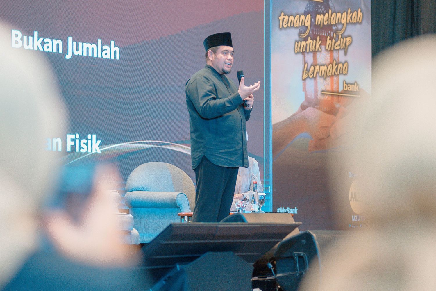 Speaker presenting on stage at a business conference with projection screen and audience in background.