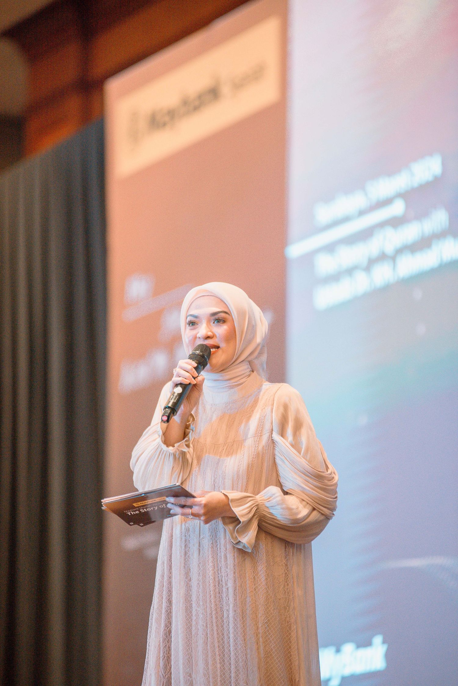 A speaker in light-colored attire gestures while presenting on stage with a projected screen in the background.
