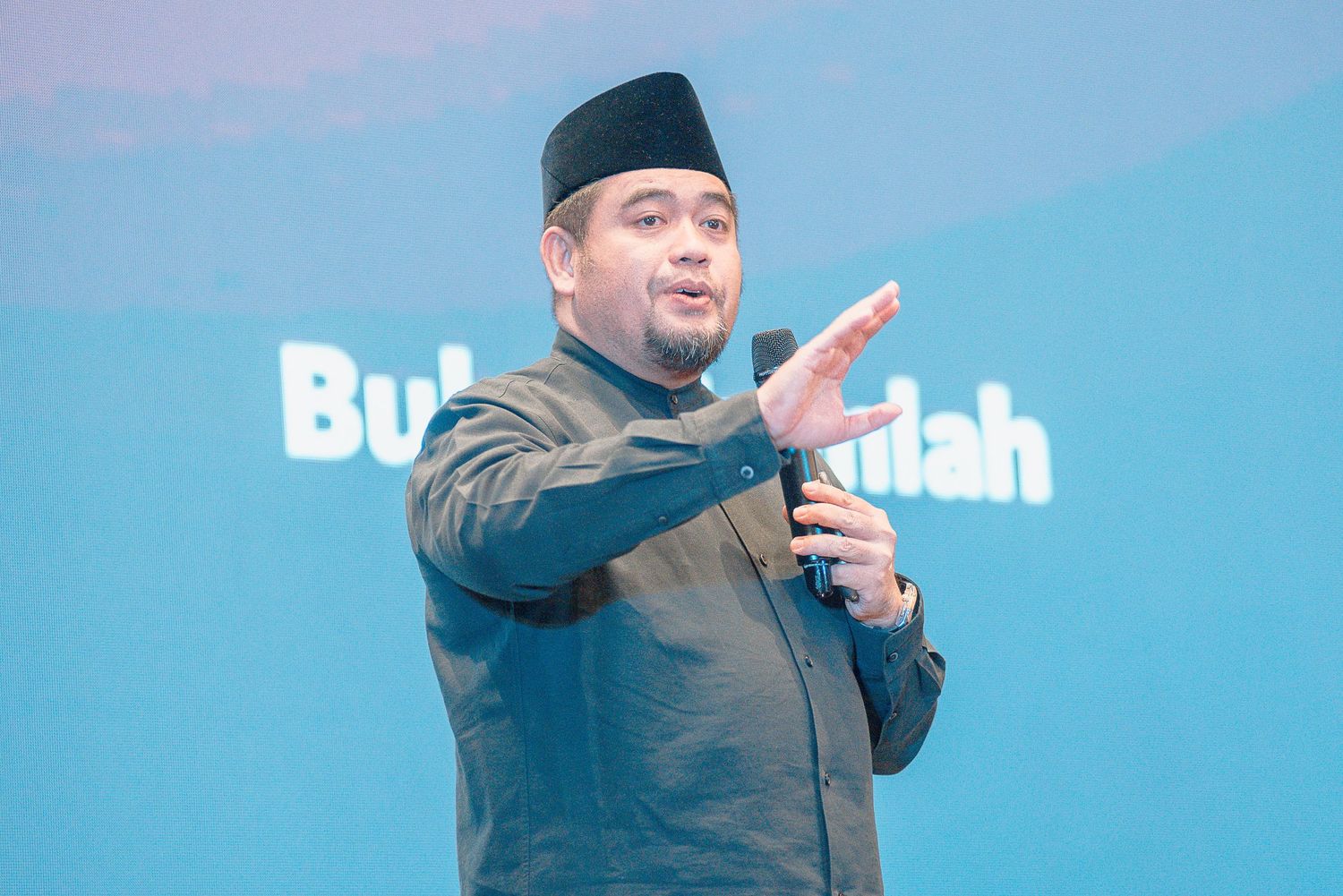 Person in black cap and jacket gestures while speaking against blue background during presentation.