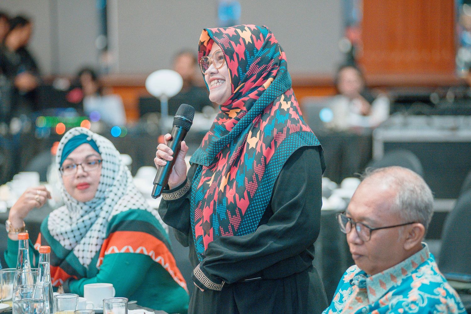A group of people wearing colorful hijabs and head coverings sit together at tables during a gathering.