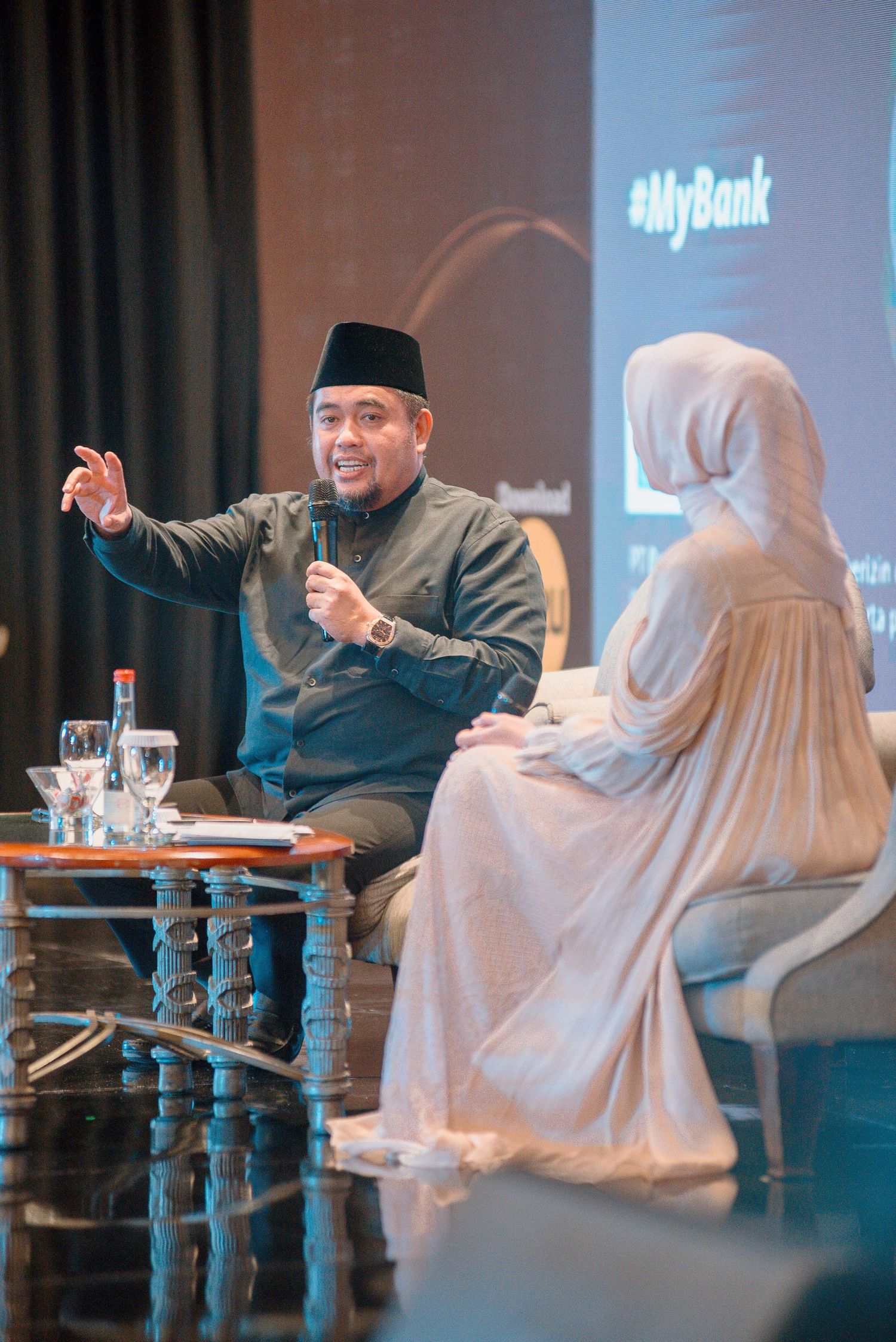Two people in traditional Muslim attire engaged in conversation at a corporate event with MyBank branding visible.