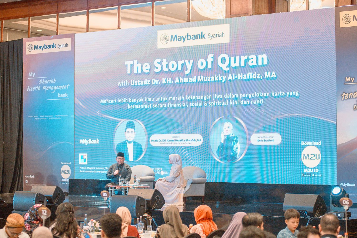Conference attendees seated in rows face a large presentation screen displaying text about The Story of Quran.