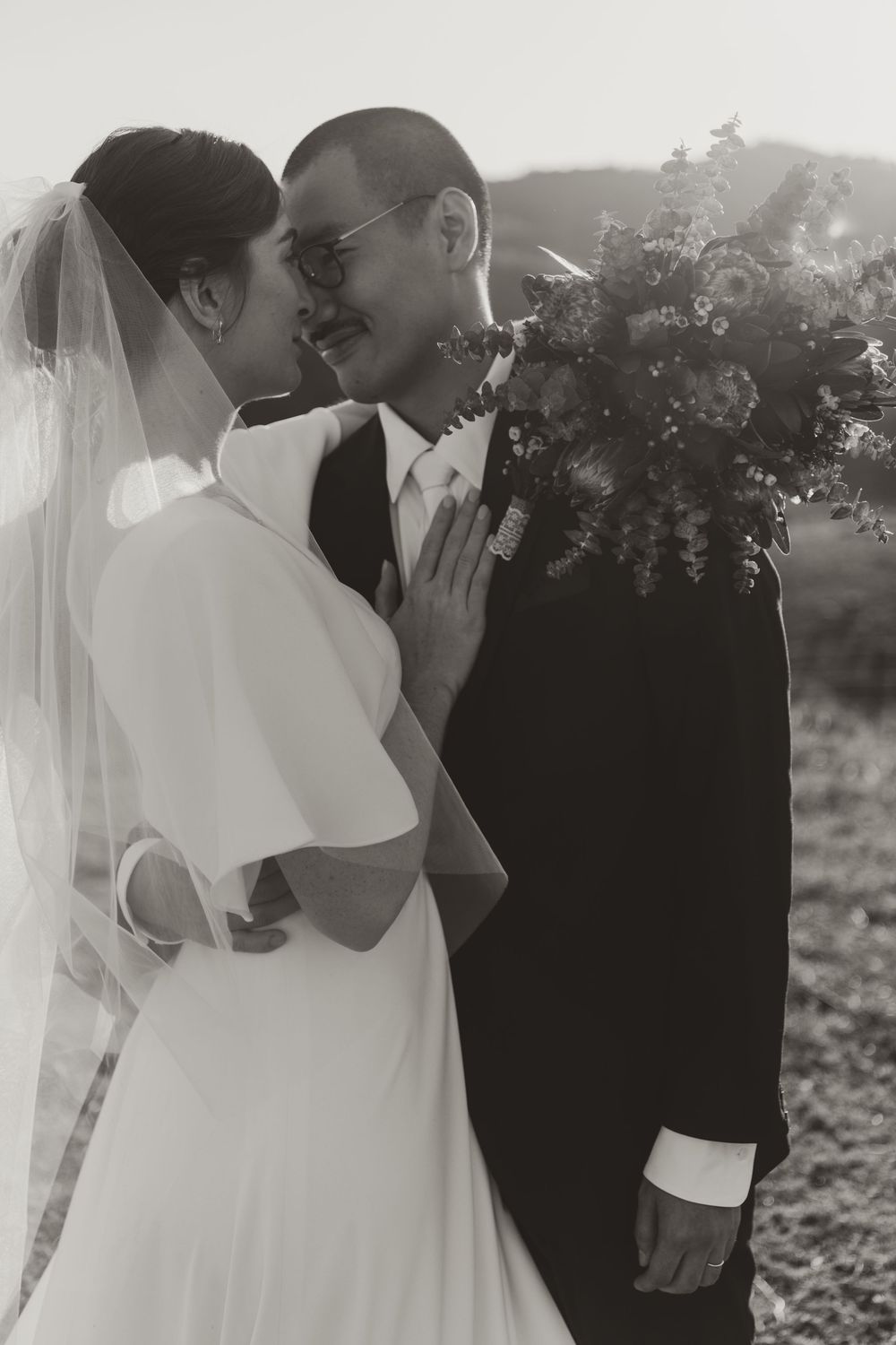Black and white wedding photo of a smiling bride in white dress and veil with groom in suit holding a floral bouquet outdoors.